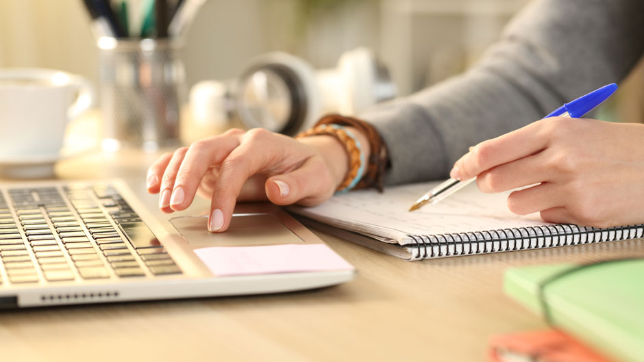 Close up of student girl hands comparing notes on notebook with laptop at home