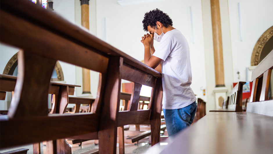 A young man kneels in prayer in an empty church pew