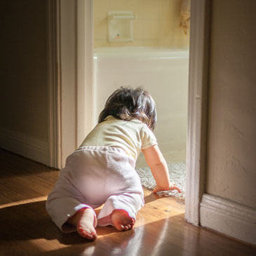 An 8 month old baby girl is crawling along hardwood floor, exploring her home. She's about to crawl into the bathroom, where light is treaming into the dark passage.
