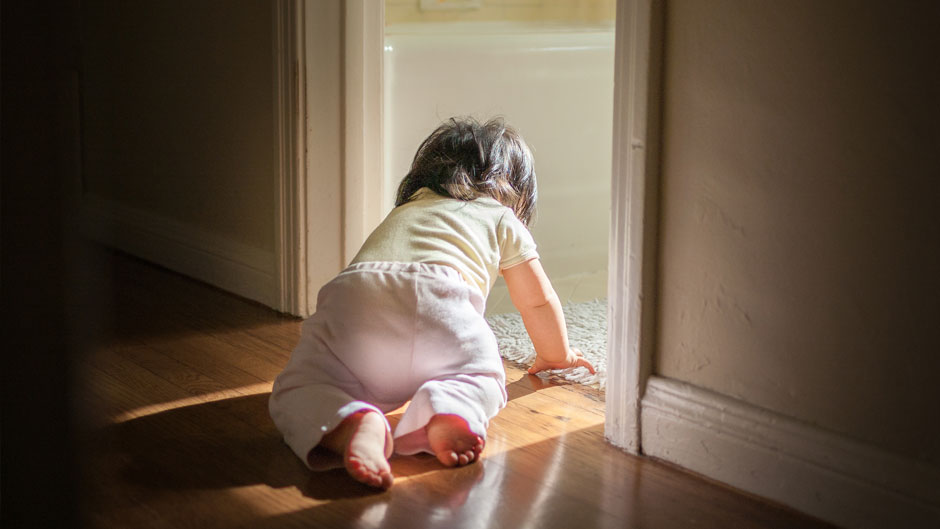 An 8 month old baby girl is crawling along hardwood floor, exploring her home. She's about to crawl into the bathroom, where light is treaming into the dark passage.