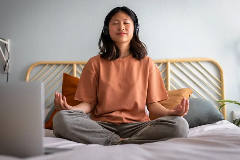 Student sitting on bed wearing headphones while meditating. 