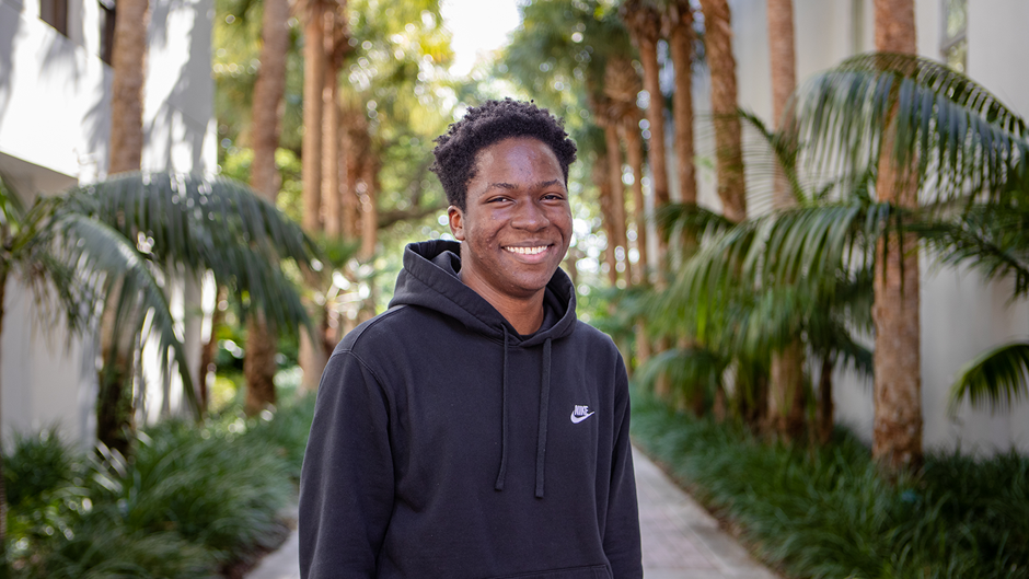 A black male student in a black Adidas hoodie smiles, standing in an outdoor courtyard. Palm trees line the path behind him.