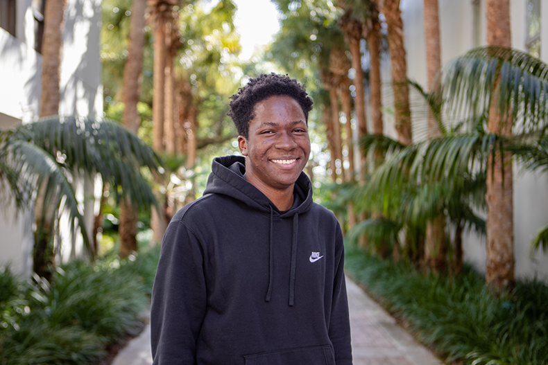 A black male student in a black Adidas hoodie smiles, standing in an outdoor courtyard. Palm trees line the path behind him.
