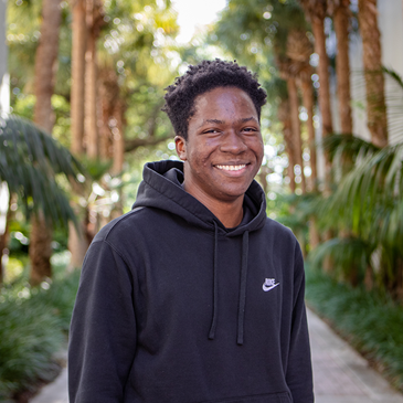 A black male student in a black Adidas hoodie smiles, standing in an outdoor courtyard. Palm trees line the path behind him.